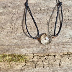 May include: A clear quartz crystal pendant on a black cord necklace. The crystal is round and encased in copper wire. The necklace is displayed on a weathered wooden surface.