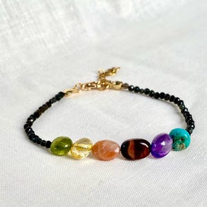 Peut inclure: Bracelet composé d'une chaîne dorée et d'une rangée de petites perles noires. Il présente sept pierres précieuses ovales de couleurs verte, jaune, orange, marron, violet et turquoise. Le bracelet est posé sur une surface blanche.