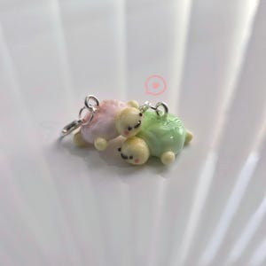 Op de afbeelding: Een paar miniatuur schildpadbedels in pastelroze en groen. Elke schildpad heeft een crèmekleurig hoofd en kleine zwarte ogen. De bedels zijn bevestigd aan een zilverkleurige sluiting, perfect voor een sleutelhanger of sieraad.