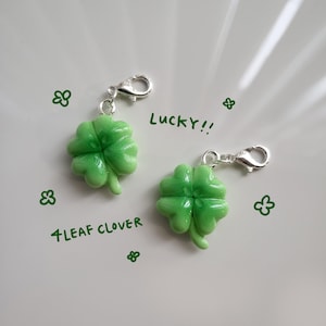 Può includere: Due ciondoli d'argento con ciondoli a forma di quadrifoglio. I ciondoli sono verdi e hanno una finitura lucida e lucida. Il testo "Lucky!!" e "4 Leaf Clover" è scritto sullo sfondo bianco.