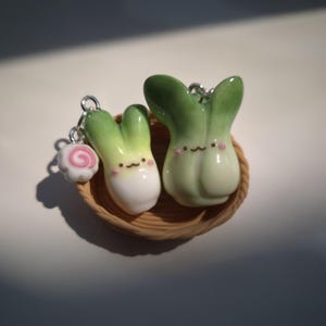 May include: Two small, glossy, vegetable-shaped charms with smiling faces and pink cheeks, one resembling a leek and the other a bok choy, resting in a miniature woven basket. A tiny white and pink narutomaki fish cake charm is attached to the leek.