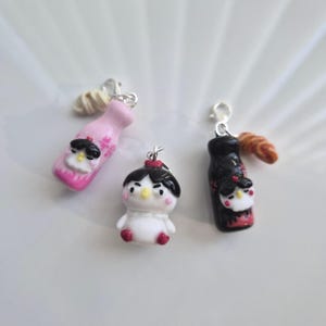 Pode incluir: Três amuletos de argila polimérica feitos à mão. Um é uma pequena figura branca com um coque preto e um laço vermelho. Os outros dois têm formato de garrafa, um rosa e outro preto, cada um com um design de rosto semelhante. Todos têm fechos prateados.