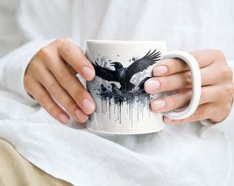 Puede incluir: Taza de cerámica blanca con un gráfico en blanco y negro de cuervos en vuelo. Los cuervos están representados en un estilo de acuarela con salpicaduras de tinta negra.