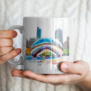 May include: White ceramic mug with a colorful watercolor illustration of the Cloud Gate sculpture in Chicago. The sculpture is a large, bean-shaped structure made of stainless steel.
