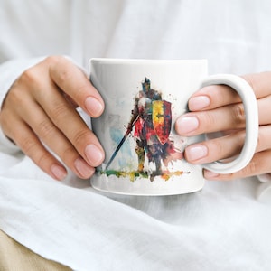May include: White ceramic mug with a watercolor illustration of a knight in armor holding a sword and shield. The knight is standing on a grassy field with a blue sky in the background.