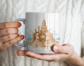 Könnte beinhalten: Eine weiße Keramiktasse mit einer Aquarell-Illustration eines Sandkastens am Strand. Das Sandkasten ist detailliert mit Türmen, Fenstern und einer Zugbrücke.