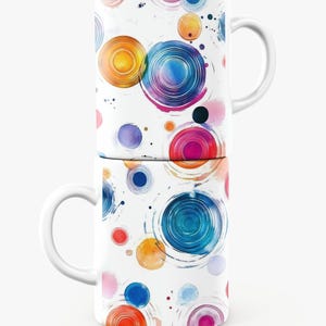 May include: Two white ceramic mugs stacked, each adorned with a vibrant watercolor design of concentric circles in shades of blue, orange, pink, and purple. The mugs have white handles and a glossy finish, creating a cheerful and artistic aesthetic.