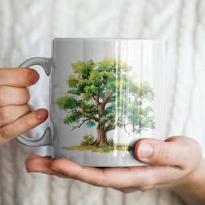 May include: A white ceramic mug featuring a watercolor painting of a large tree with green leaves and a brown trunk. The mug has a curved handle and is held by a person wearing a white sweater.