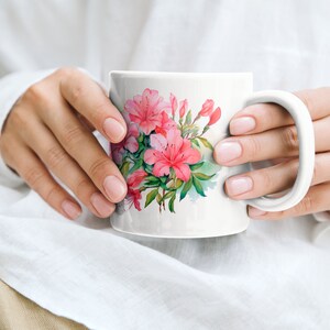 K&ouml;nnte beinhalten: Eine wei&szlig;e Keramiktasse mit einem rosafarbenen Blumendesign mit Azaleen. Die Tasse hat einen Henkel und wird von den H&auml;nden einer Person gehalten.