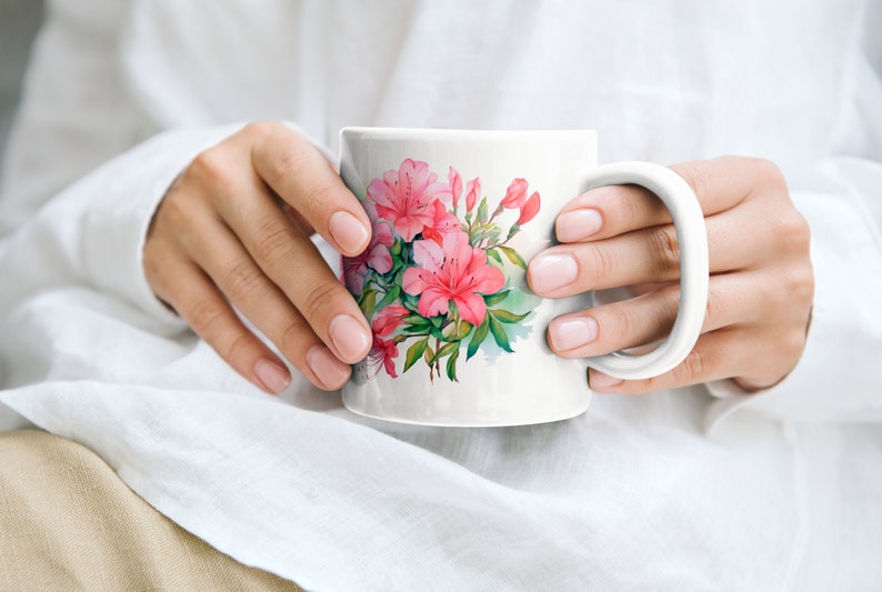 K&ouml;nnte beinhalten: Eine wei&szlig;e Keramiktasse mit einem rosafarbenen Blumendesign mit Azaleen. Die Tasse hat einen Henkel und wird von den H&auml;nden einer Person gehalten.
