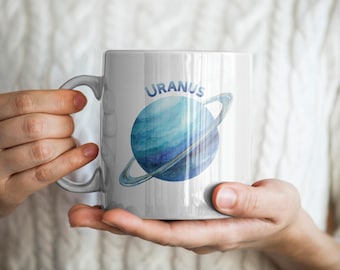 May include: White ceramic mug featuring a watercolor illustration of the planet Uranus, with blue and teal tones and a ring. The word "URANUS" is printed above the planet. The mug is held by a person.