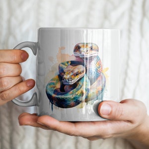 May include: White ceramic mug featuring a watercolor illustration of two colorful snakes. The snakes are depicted in shades of blue, yellow, orange, and green. The mug is held by a person wearing a white sweater.