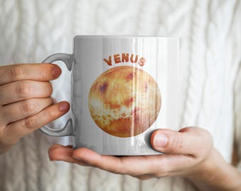 May include: White ceramic mug featuring the planet Venus, with the planet's image and the name "VENUS" in orange lettering. The mug is held by a person.
