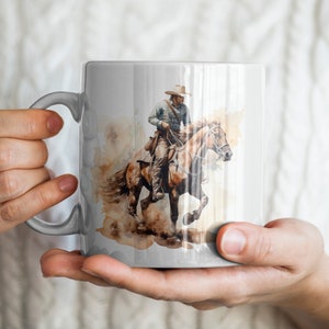 May include: A white ceramic mug with a watercolor illustration of a cowboy riding a brown horse. The cowboy is wearing a brown hat and a blue shirt. The horse is running and its mane is flowing in the wind.