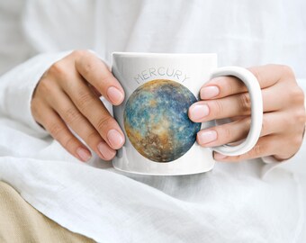 May include: White ceramic mug featuring a colorful depiction of the planet Mercury. The word "MERCURY" is printed above the planet image. The mug has a curved handle and is held by a person.