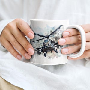 May include: A white ceramic mug with a watercolor design of a military helicopter. The helicopter is gray and black and is flying in the sky. The mug is being held by a person's hands.