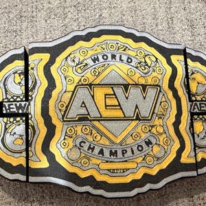 May include: A black, gold, and silver AEW World Champion wrestling belt. The belt features the AEW logo and the words "WORLD CHAMPION". The belt has a detailed design with globe and decorative elements.