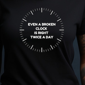 以下が含まれることがあります： 黒の半袖Tシャツ。文字盤の目盛りと中央に「EVEN A BROKEN CLOCK IS RIGHT TWICE A DAY」というテキストが白でプリントされています。デザインは衣服の胸元部分に、すっきりとしたサンセリフ体のフォントで配置されています。