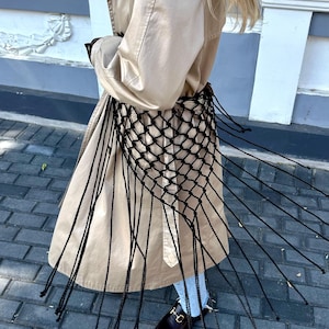 May include: A tan trench coat with a black net overlay and fringe detail. The coat has a classic design with a double-breasted front and a belted waist. The black net adds a unique texture and visual interest to the garment.
