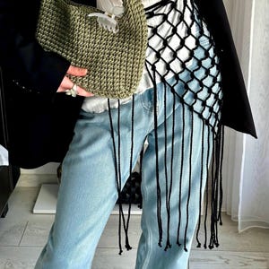 May include: A green crocheted handbag with a short handle and a decorative butterfly clip. The bag is held by a person wearing light blue jeans, a white top, and a black blazer. A black net-like garment with long fringe hangs over the jeans.