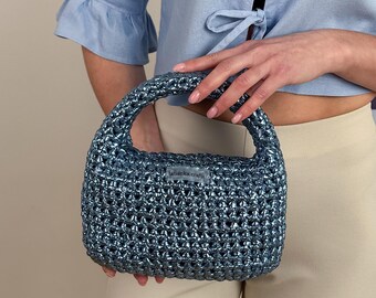 May include: A small, light blue crocheted handbag with a silver sheen. The bag has a single top handle and a small label that reads "tatianka crafts".