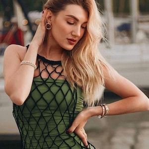 May include: A woman wearing a green and black fishnet dress with a halter neckline. She is standing in front of a blurred background.