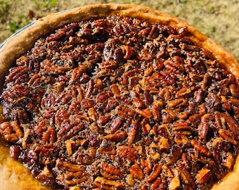 Könnte beinhalten: Ein ganzer Pekannusskuchen in einer Alufolie. Der Kuchen hat einen goldbraunen Boden und ist mit einer glänzenden, dunkelbraunen Pekannussfüllung gefüllt. Der Kuchen steht auf einer Oberfläche mit grünem Gras im Hintergrund.