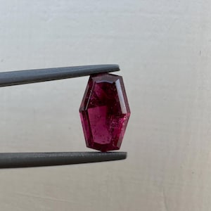 以下が含まれることがあります： ファセットカットされた八角形の濃いピンクの宝石。宝石はピンセットで保持されています。