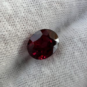 Puede incluir: Una piedra preciosa ovalada de color rojo intenso con facetas, sobre un fondo de tela blanca.