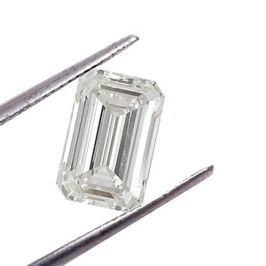 May include: A clear, emerald-cut diamond held by silver tweezers against a white backdrop. The diamond's facets reflect light, highlighting its clarity and shape. The rectangular cut is a classic for jewellery.