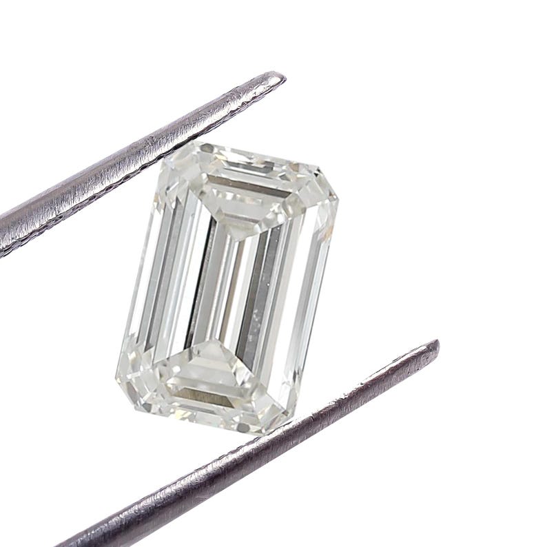 May include: A clear, emerald-cut diamond held by silver tweezers against a white backdrop. The diamond's facets reflect light, highlighting its clarity and shape. The rectangular cut is a classic for jewellery.