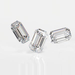 May include: Three clear emerald-cut diamonds are shown against a white backdrop. The diamonds have parallel step cuts, forming a rectangular shape with cut corners. The facets reflect light, creating a brilliant sparkle.