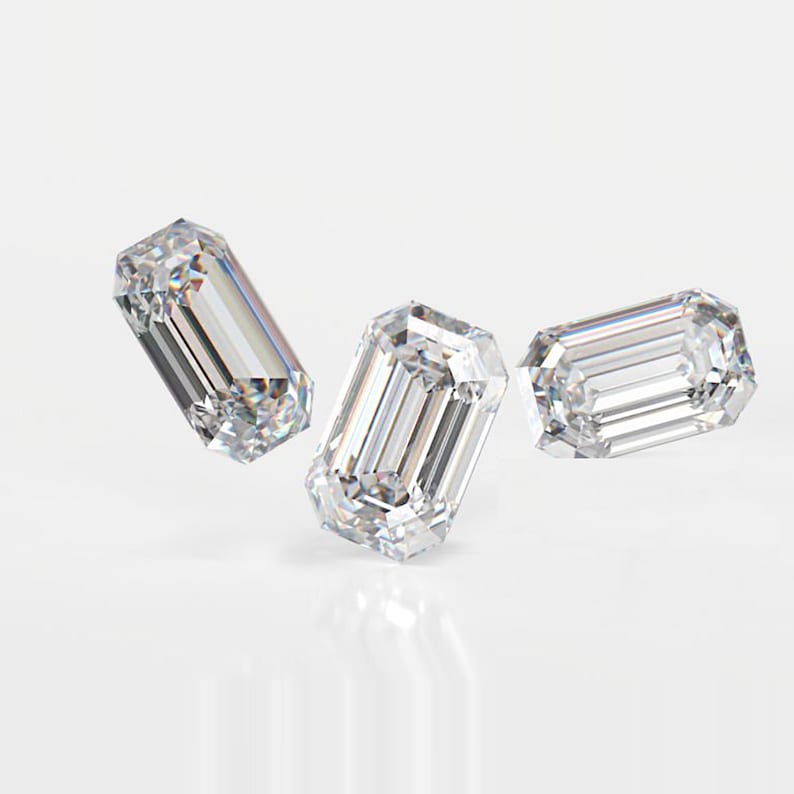 May include: Three clear emerald-cut diamonds are shown against a white backdrop. The diamonds have parallel step cuts, forming a rectangular shape with cut corners. The facets reflect light, creating a brilliant sparkle.