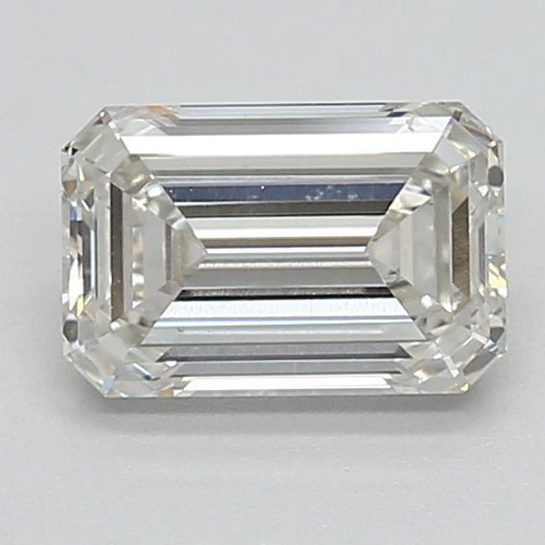 May include: A clear, emerald-cut diamond. The rectangular gemstone features step-cut facets, creating a brilliant display of light. The diamond's clarity is high, and it is set against a neutral background to enhance its sparkle and brilliance.