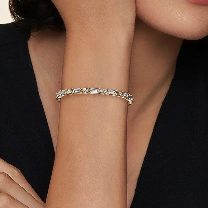 Op de afbeelding: Een gouden armband met afwisselend rechthoekige en ronde heldere stenen. De armband wordt om een pols gedragen, met een zwarte top op de achtergrond. De armband is een sieraad.