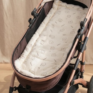 May include: A baby stroller featuring a rose gold frame and a dark brown seat unit. Inside the seat is a cream-colored padded liner with a repeating pattern of small brown line-art rabbit faces and the word Rabbit printed in a simple font.