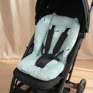 May include: A light mint green stroller liner featuring a repeating pattern of white polar bears wearing green scarves and white snowflakes. The liner is installed in a black stroller with a five-point black harness system and a black canopy.