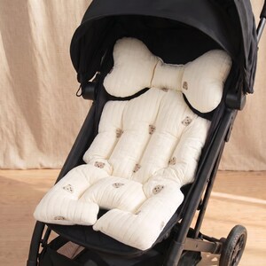 May include: A black stroller featuring a cream-colored padded seat liner with a vertical quilted texture and a repeating pattern of small brown bear faces. The liner includes a contoured headrest section and integrated side support cushions.