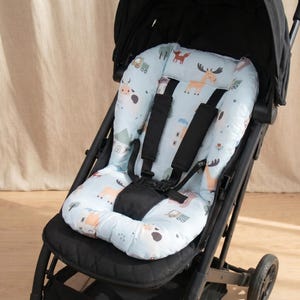 May include: A light blue stroller seat liner featuring a repeating pattern of cartoon animals including moose, cows, and foxes, along with small trucks and houses. The liner includes black harness straps and a central buckle for secure attachment.