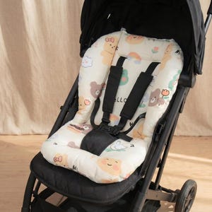 May include: A black stroller featuring a cushioned seat liner with a cream-colored fabric cover. The liner is decorated with a repeating pattern of cartoon bears, rainbows, clouds, and flowers. It includes a black five-point safety harness system.