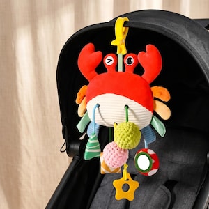 Pode incluir: Um brinquedo de pelúcia em forma de caranguejo com topo vermelho brilhante e base de veludo cotelê creme. Possui acessórios pendurados, incluindo esferas texturizadas, um pequeno espelho e um anel amarelo em forma de estrela com clipe.