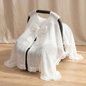 May include: A white muslin car seat cover featuring a textured, lightweight fabric with a ruffled hem and a large decorative bow centered on the front. The cover is draped over a black plastic infant car seat, providing a soft, semi-sheer layer.