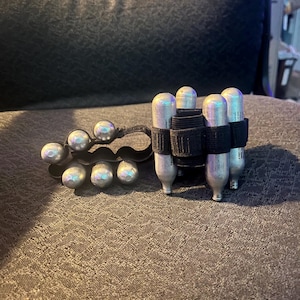 May include: A black paintball pod holder with silver pods and a black wrist strap. The image also shows a black holder with silver pods.