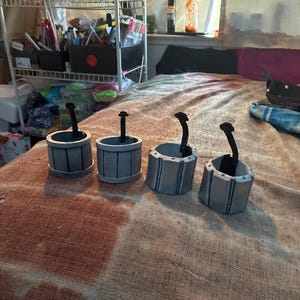 May include: Four cylindrical silver-colored containers with vertical black lines and a single black handle extending upward from the center of each. The containers feature a metallic finish and a structured design, arranged in a row on a textured surface.