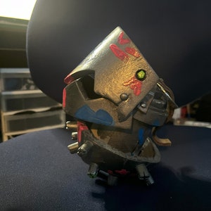 May include: A small, metallic, multi-colored robot figurine with a green eye and red and blue accents. The robot is made of multiple pieces and has a textured surface.