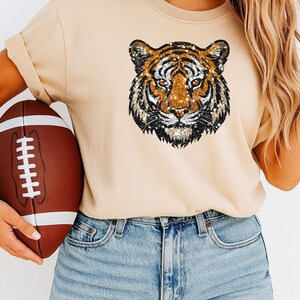 May include: A tan-colored sweatshirt featuring a sequined tiger face design. The image also includes a brown and white football. The sweatshirt is paired with blue denim jeans.