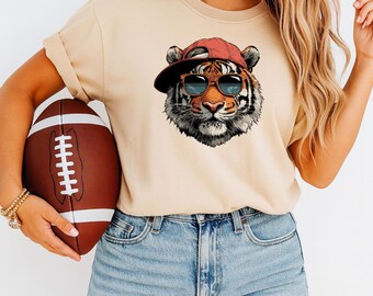 May include: A beige t-shirt featuring a tiger wearing sunglasses and a red cap. The person in the image is holding a brown football. The shirt has a crew neck and short sleeves. The person is wearing blue denim shorts.