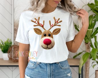 May include: White t-shirt featuring a detailed embroidered reindeer face with brown antlers and a red nose. The reindeer design is centered on the shirt. The text "PINK HONEY png" is visible in the lower left corner. The model is wearing blue denim shorts.