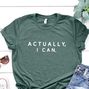 Może przedstawiać: Zielony T-shirt z napisem "Actually, I can." w białych literach. Koszulka leży płasko na białym drewnianym tle z okularami przeciwsłonecznymi, białymi butami sportowymi i niebieskimi dżinsami.