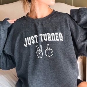 Puede incluir: Una sudadera gris oscuro con el texto "JUST TURNED" y dos gestos con las manos, un signo de paz y un dedo medio, impresos en blanco.